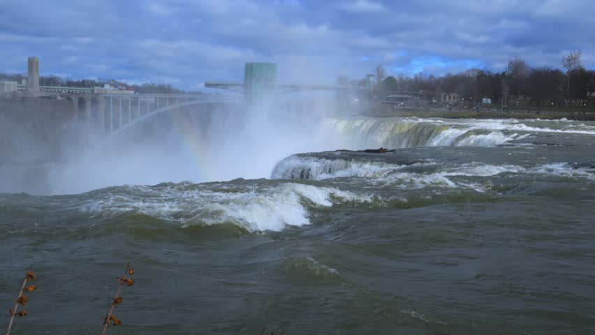 Niagara Falls, USA - November 26, 2025: This video captures the Niagara Falls from the U.S. side, featuring the rushing Niagara River in the foreground. In the background, the iconic Rainbow Bridge connects the American side to Canada. The turbulent water depicted is consistent with the Bridal Veil Falls and American Falls area.
