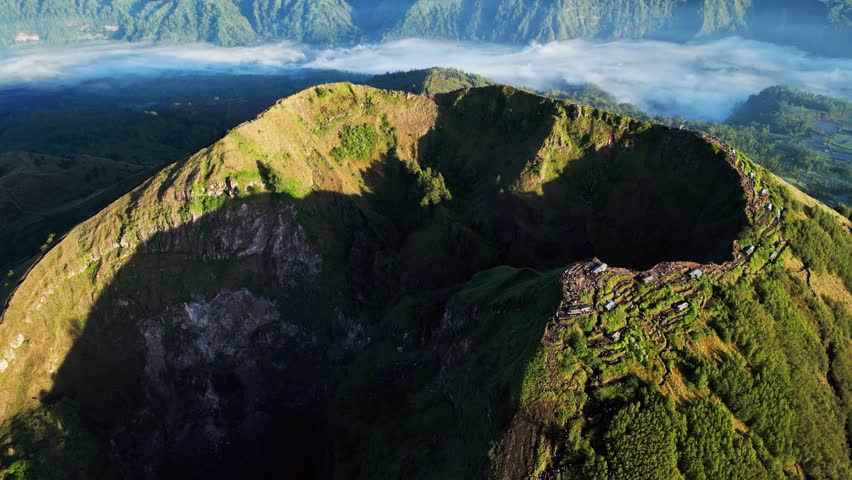 Volcanic crater landscape glowing in warm sunrise light with steep Mount Batur terrain, forested slopes and drifting fog across tropical Bali highlands push-in aerial drone footage.