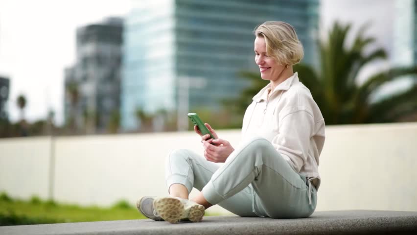 Mature blonde woman sitting comfortably outdoors holding knees and talking on phone in urban area