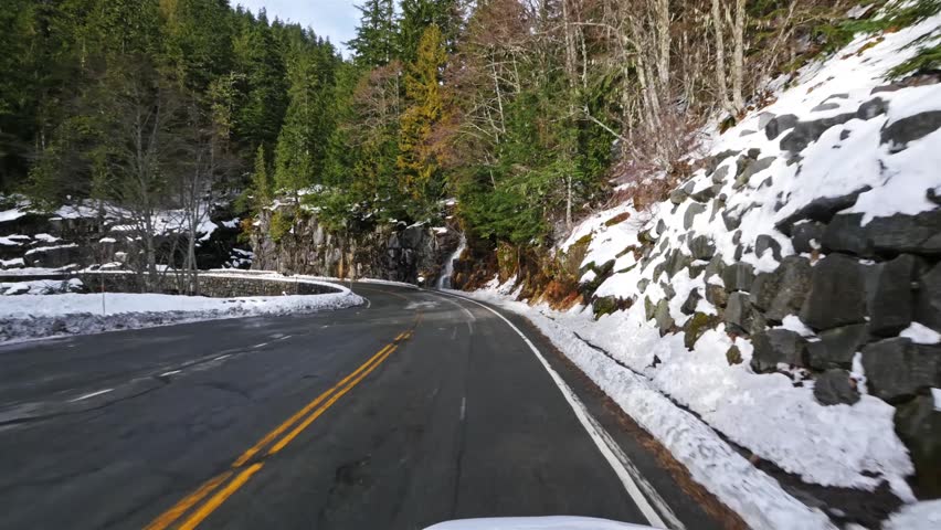 Scenic drive footage of a winding asphalt road meandering through a snow-covered forest landscape under a clear sky in Mount Rainier National Park, Washington State, USA.