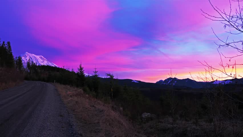 A vibrant sunset or sunrise sky featuring vivid pinks, purples, and blues over the snow-capped peak of Mount Rainier, as seen from the scenic Mowich Lake Road in Mount Rainier National Park, Washingto