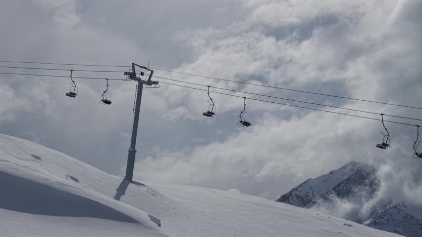 Chairlift crossing snowy alpine slopes at Las Leñas ski resort in Mendoza, Argentina, surrounded by Andes peaks, winter clouds and mountain terrain, drone lateral slide right shot.