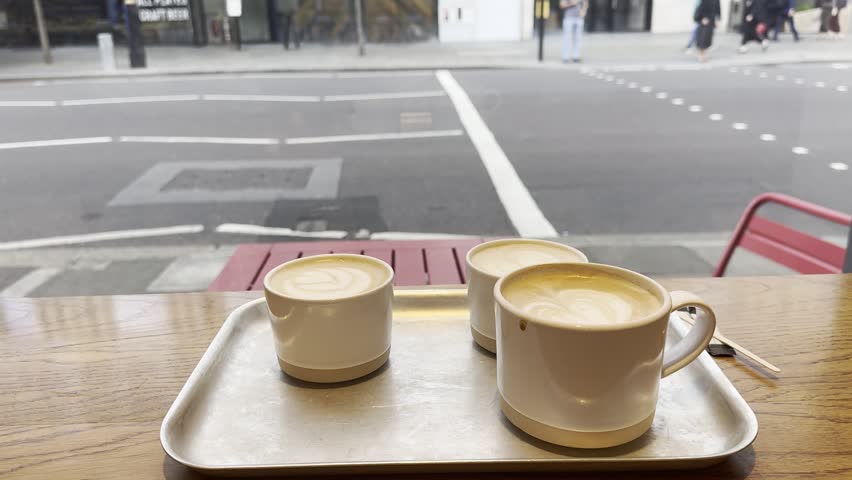 Three cups of hot cappuccino coffee stand on a tray near a window overlooking busy traffic on Buckingham Palace Road in London.