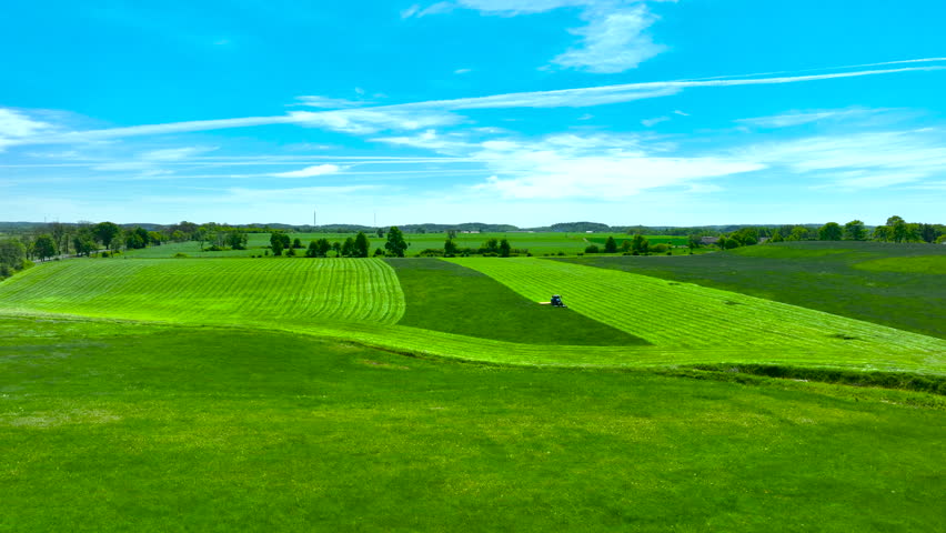 Cinematic drone shot of a tractor mowing vibrant green fields in Poland under a blue sky.