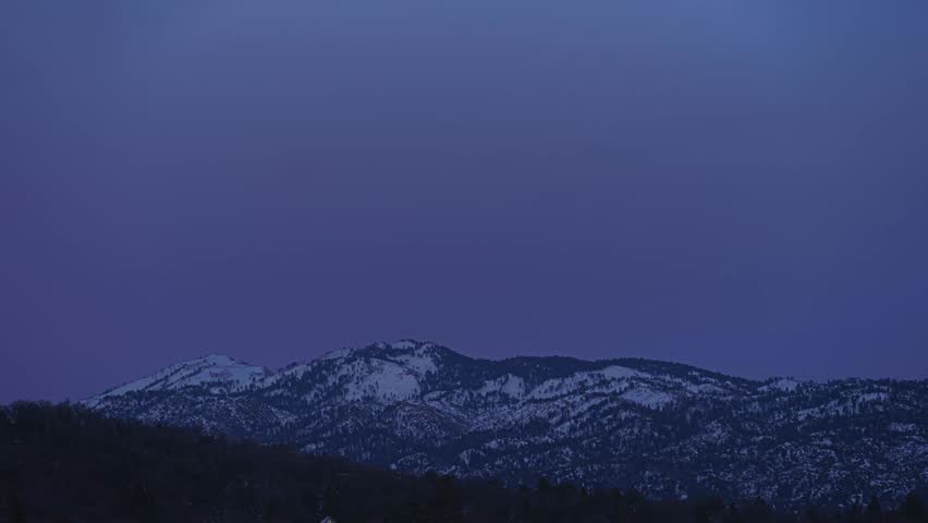 Time lapse of a yellow full moon rising over a snowy mountain ridge against a pink sky at sunset in the San Bernardino National Forest, California, filmed from Lake Arrowhead, California.