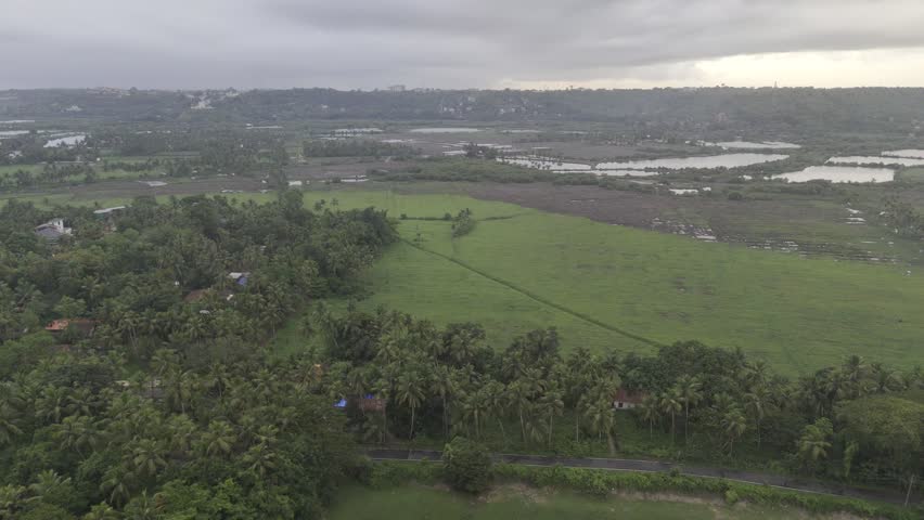 Aerial landscape view of Talaulim fields during the monsoon season, featuring lush greenery, coconut palm trees, tranquil backwaters, and a dramatic sunset glowing behind dense clouds in Goa, India.