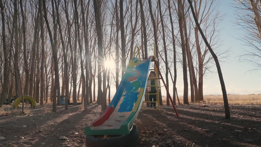 Young girl enjoying a playground slide among tall trees in warm golden sunset light.