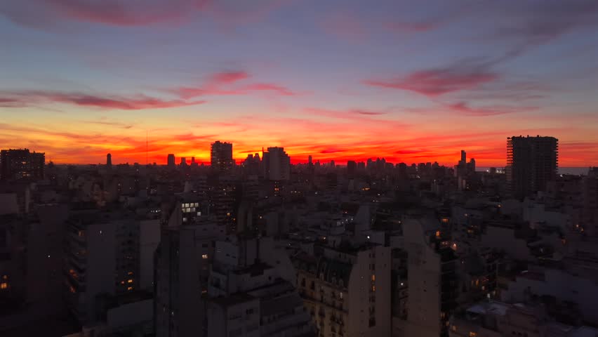 The camera moves over darkened city buildings towards a vibrant red and orange sky.