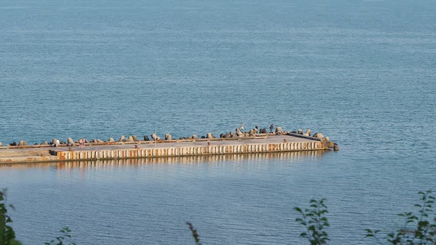 Concrete rusty pier extending into a calm blue sea, providing a resting place for a flock of seabirds while people relax in the distance, creating a serene and picturesque coastal scene