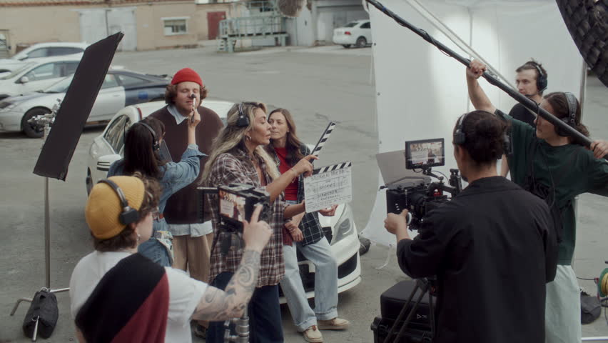 Professional film crew capturing scene with two actors sitting on car hood, smiling and chatting. Stylist applying makeup and assistant using clapperboard on urban movie set