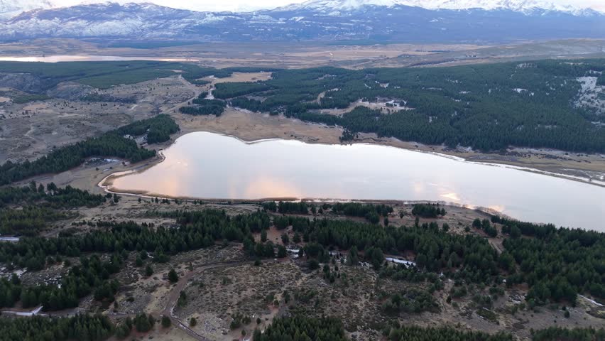 An aerial flight moves over pine forests toward a calm lagoon reflecting sunset sky.