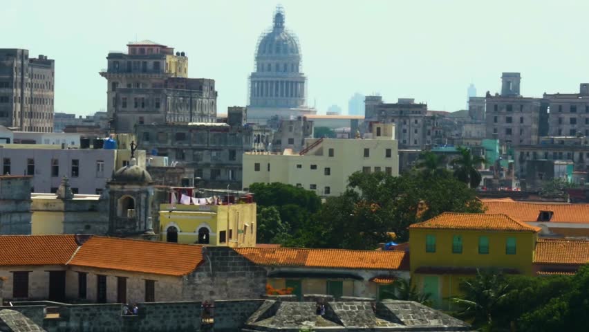 Aerial perspective slowly moving across the weathered rooftops and colorful buildings of old Havana, showing the iconic dome of El Capitolio Nacional against the hazy skyline on a sunny day