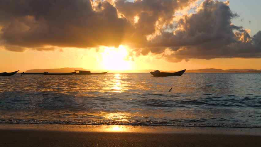 SILHOUETTE, LENS FLARE: Golden evening light pours across shoreline as boats drift on calm ocean water under dramatic clouds. Reflections shimmer over wet sand and small waves during a tropical sunset