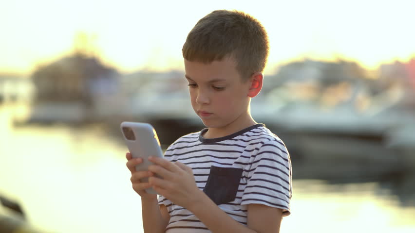 An eight-year-old boy sits on the shore and plays the phone against the backdrop of yachts and boats