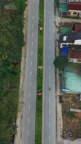 Vertical screen Aerial View Curved Village Road Phong Nha Midday, Winding Asphalt Lanes With Planted Median, Colorful Rooftops Hugging Bend, Motorbikes Weaving Past Parked Cars, Delivery