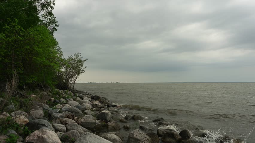 View along a rocky lakeshore with waves breaking on the rocks and trees with leaves blowing in the wind