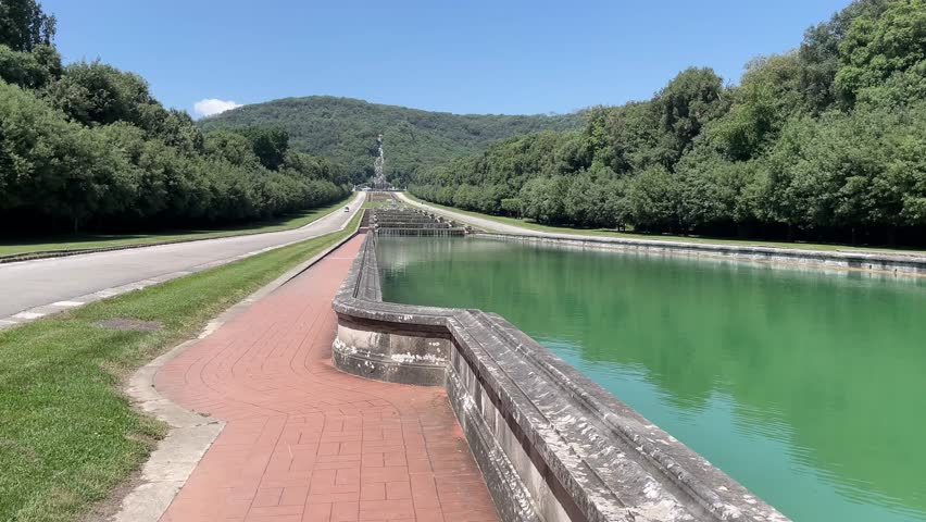 Steady static shot of a long reflecting pool and paved walkway at the Reggia di Caserta gardens, featuring landscaped tree lines and distant tiered fountains