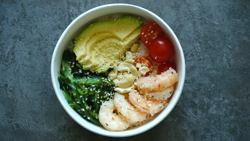 Fresh and healthy poke bowl on dark table, featuring shrimp, avocado, seaweed salad, cherry tomatoes, and cashews, representing balanced and nutritious meal. Healthy poke bowl with shrimp and avocado
