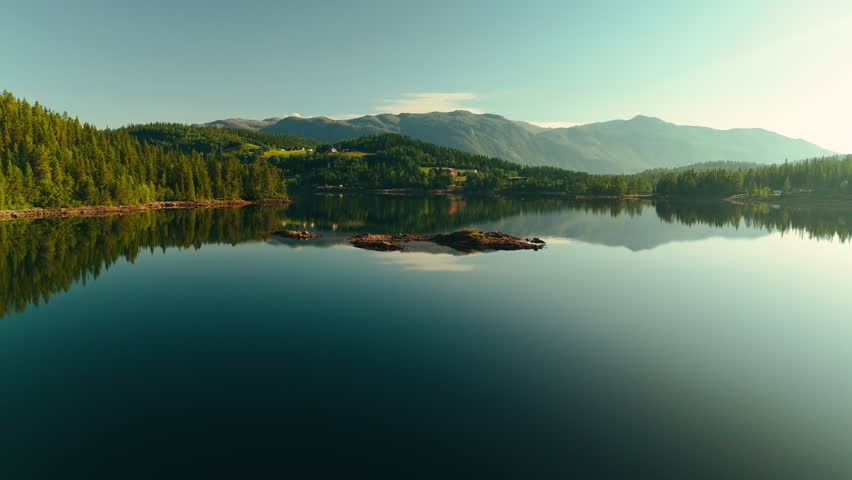 Aerial drone footage with a tranquil sunrise over a mirror-smooth lake in Norway. The calm water perfectly reflects the surrounding mountains and soft morning light