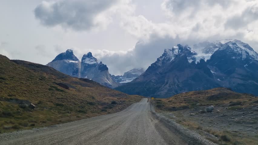 POV passenger view from a moving car approaching the Cuernos del Paine in Torres del Paine National Park, Chile, with dramatic peaks rising ahead under dark Patagonian clouds
