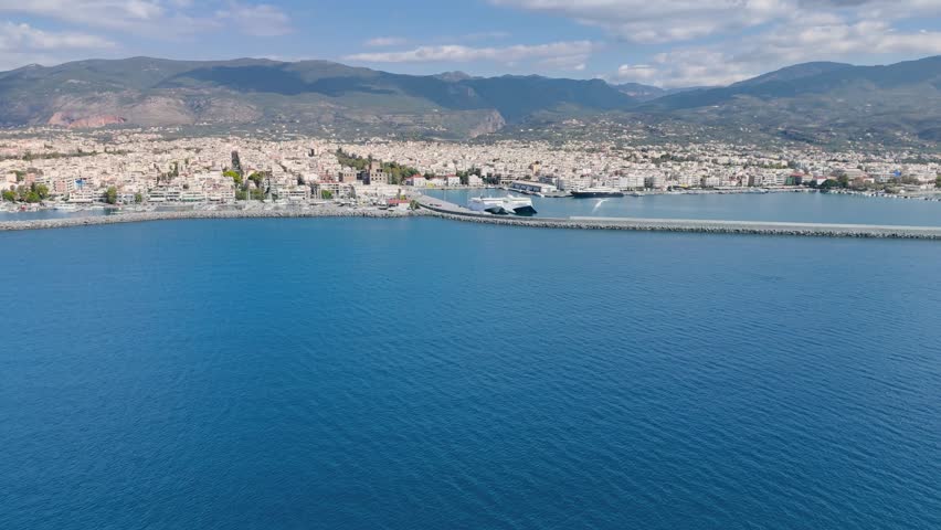 Aerial drone footage capturing the harbor district of Kalamata, one of the largest coastal cities in the Peloponnese region of Greece. The video showcases the waterfront, marina, breakwater, ferries, port facilities, and dense urban cityscape stretching along the shoreline. The Taygetus Mountains create a dramatic natural backdrop while the calm waters of the Messinian Gulf provide a striking Mediterranean setting. Suitable for travel documentaries, tourism promotion, maritime transportation content, city destination videos, Greek geography projects, infrastructure presentations, and coastal urban establishing shots.