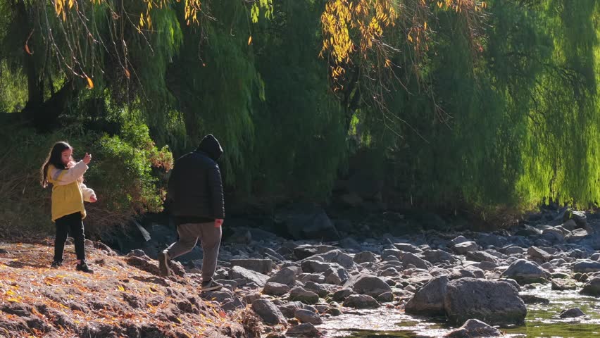 Father and daughter exploring rocky lakeshore beneath green willow trees, pan across riverbed