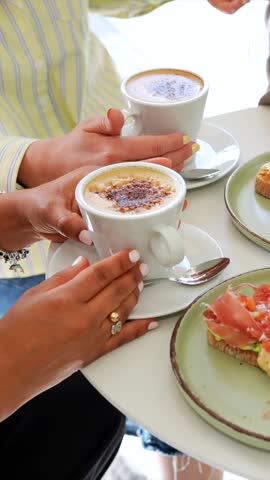 Two women enjoying a delicious brunch at a coffee shop with cups of cappuccino and various toasts, including avocado with prosciutto, banana with nuts, and tomatoes with arugula