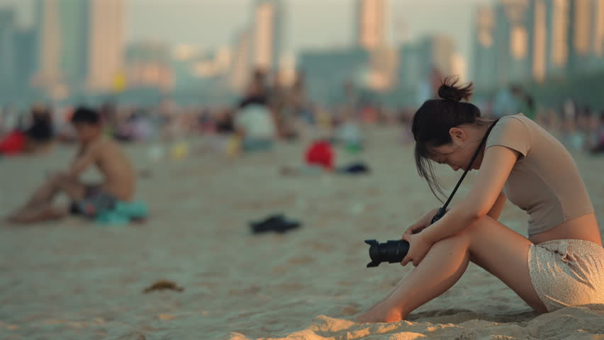 Young female photographer holding camera on crowded beach during sunset in Da Nang Vietnam, Asian woman enjoying seaside photography and travel lifestyle on sandy coastline at golden hour 4k