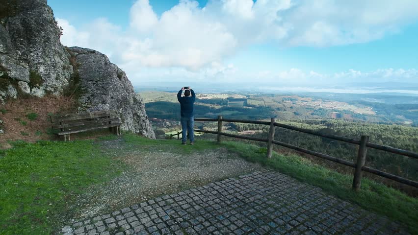 Solitary man enjoying a stunning mountain top view at a scenic lookout in Galicia, Spain, taking a picture with his smartphone before walking towards the camera on a bright and cloudy day