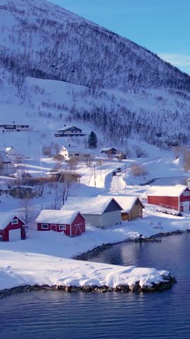 Snow covered fishing village on coastline in winter, Norway. Surroundings of town Tromso. Panoramic aerial view landscape of nordic mountains, houses, rorbu, boats and ocean. Troms county, Fjordgard