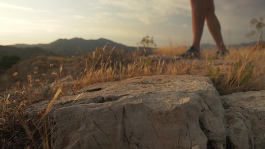 A person wearing barefoot shoes steps forward onto a large rock on a dry grassy hillside at golden hour sunset, surrounded by serene natural scenery.