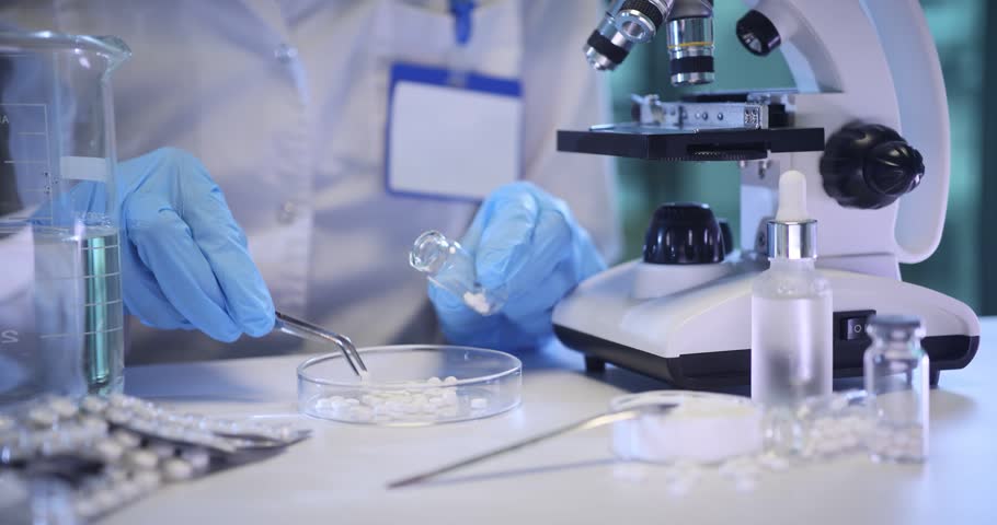 Pharmacist hands fill glass bottle with small pills at table in lab. Medical worker packs experimental supplement by microscope in chemical laboratory