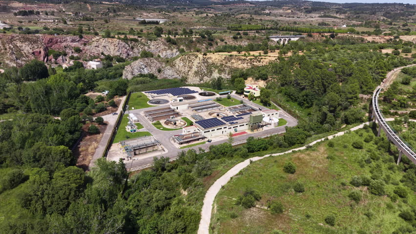 Wastewater treatment plant with solar panels nestled among green hills in Spain, featuring circular settling tanks, industrial buildings and surrounding Mediterranean landscape