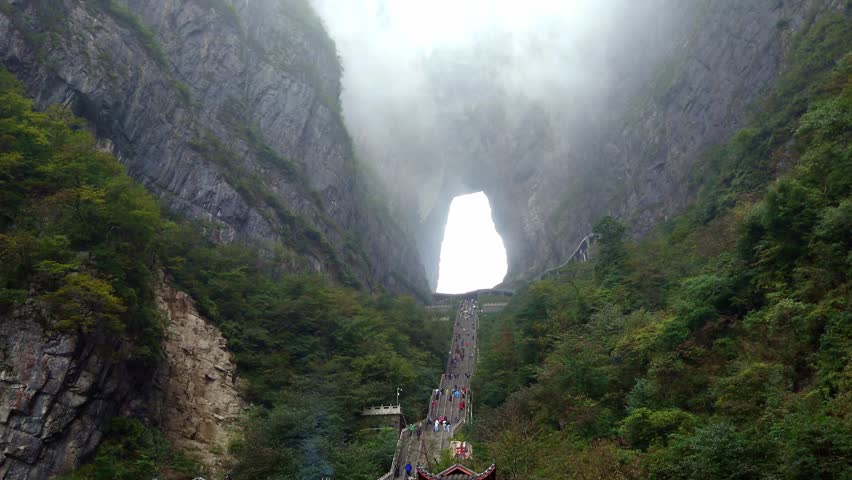 A dramatic low-angle shot of steep 999 steps staircase and the massive natural archway of Tianmen Cave on Tianmen Mountain. Thick, moody mountain fog rolls through the giant stone aperture.