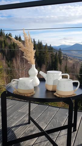 The wind shakes the branches in a vase standing on a table. Stylish ceramic tea set on the terrace with a view of the Carpathian Mountains. Aesthetic relaxation, a cozy morning, coffee, tea