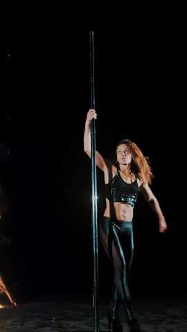 Pole dance. witch halloween. fit female gymnast, in black leather outfit and high heels, performs acrobatic exercises on metal rotating pole, at night, by light of large fire in background.