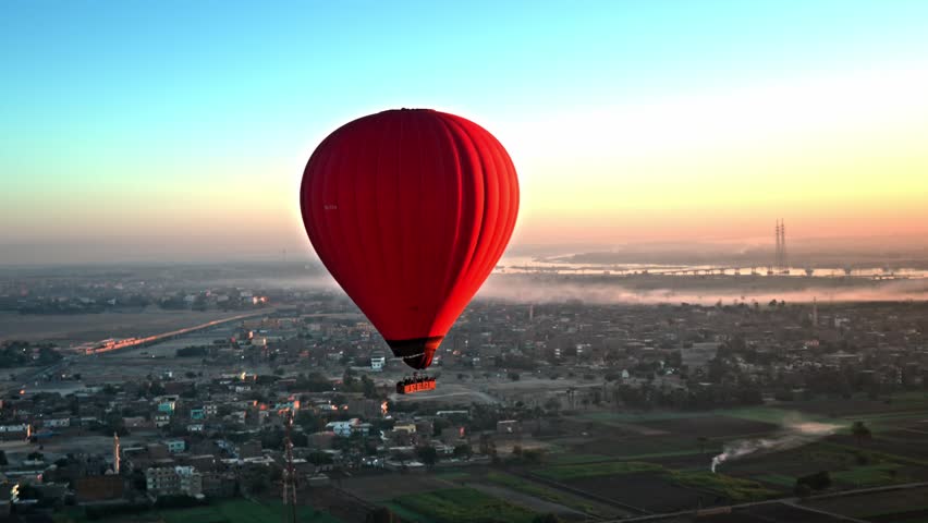 A stunning aerial view featuring a vibrant red hot air balloon drifting over the West Bank of Luxor at dawn, with the Nile River, hazy city landscape, and the soft golden glow of sunrise.