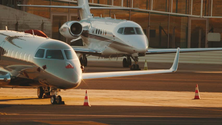 Two private jets are parked on the tarmac of an airport as the sun sets in the background. The runway is busy with activity as staff prepare for upcoming flights