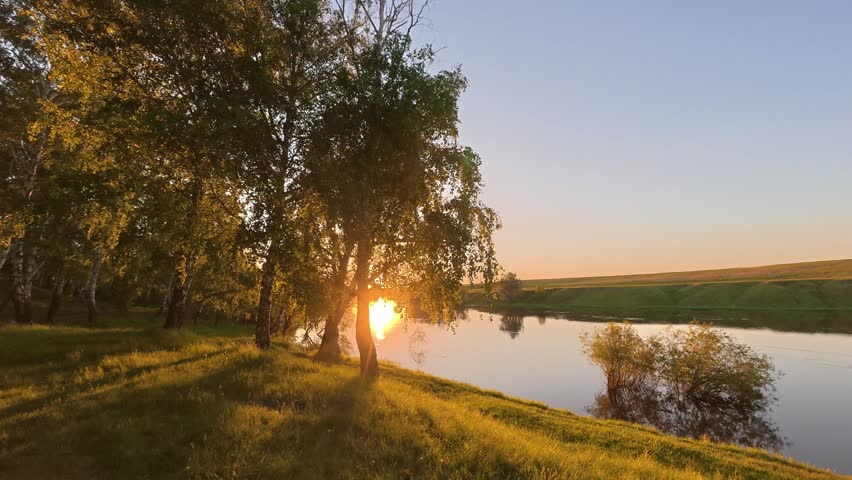 A beautiful time lapse captures the sun setting over a calm river with birch trees on the bank