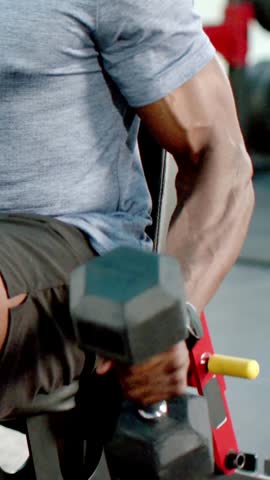 Vertical video: Gripping hex dumbbell, man in tee pressing up on incline bench, training shoulders. Gym, weightlifting, weights, strength, fitness, athlete, vertical video