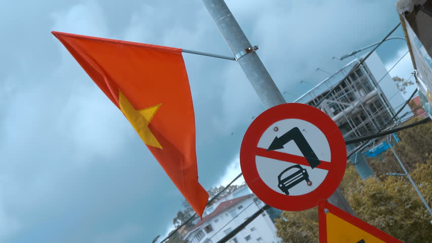 A waving flag of Vietnam and a traffic sign indicating a restriction on vehicular movement on a street pole in a public space in Hanoi City
