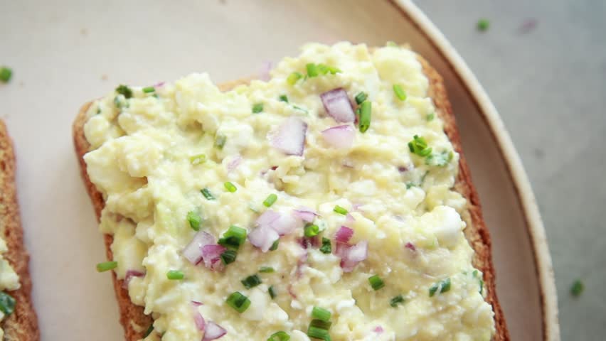 Close-up of a person spreading a creamy mixture of avocado, hard-boiled egg, and cottage cheese onto a slice of toasted whole grain bread