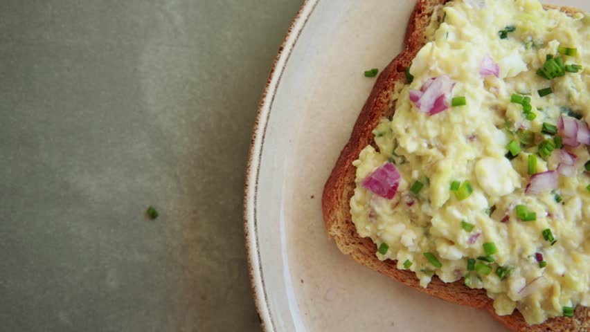 Close-up of a person spreading a creamy mixture of avocado, hard-boiled egg, and cottage cheese onto a slice of toasted whole grain bread