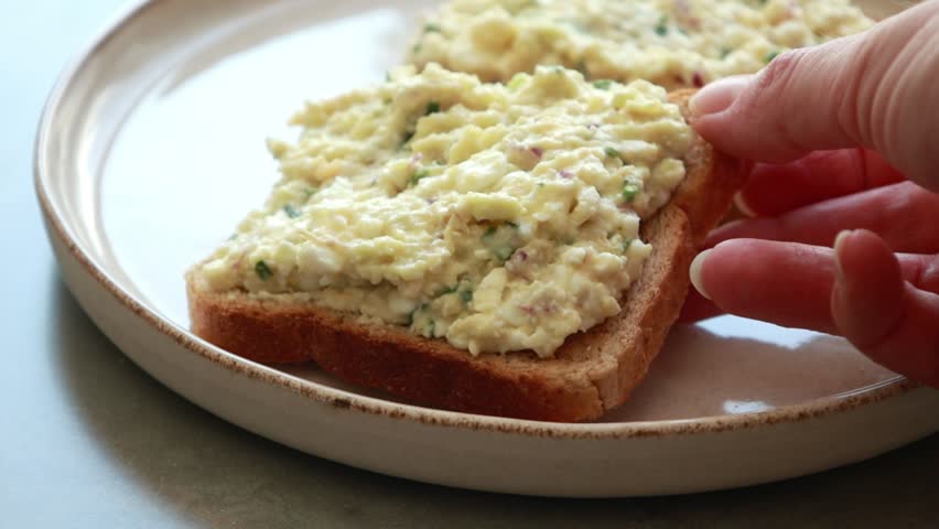 Close-up of a person spreading a creamy mixture of avocado, hard-boiled egg, and cottage cheese onto a slice of toasted whole grain bread
