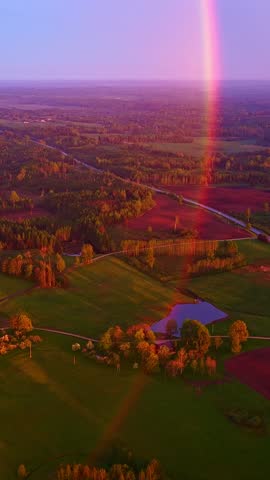 A scenic drone shot shows a beautiful rainbow touching down across green fields, forests, and a small pond.