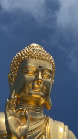 Vertical view shows golden Buddha statue seated calmly against bright cloud background. Tranquil spirituality.