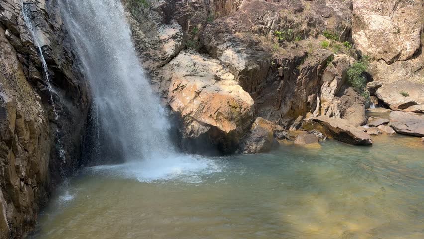 A waterfall stream drops into a natural rock basin at a cliff base forming a quiet freshwater cascade among rugged stone formations. Mountain waterfall.