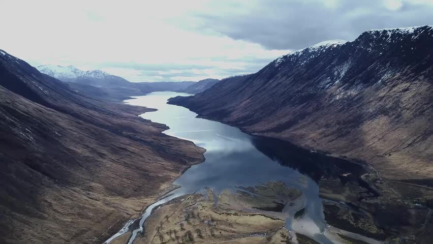 A spectacular high-altitude drone shot overlooking the Scottish Highlands, featuring a winding loch stretching through a dramatic mountain landscape. The expansive aerial perspective reveals rugged pe