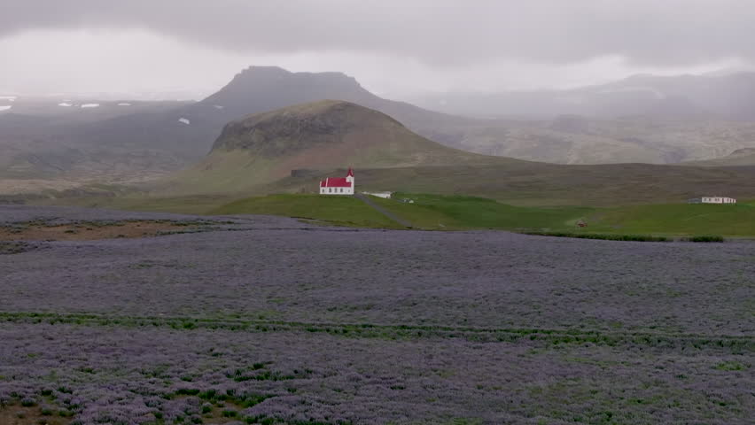 Wide aerial pan of rural Icelandic church with green fields and distant mountains