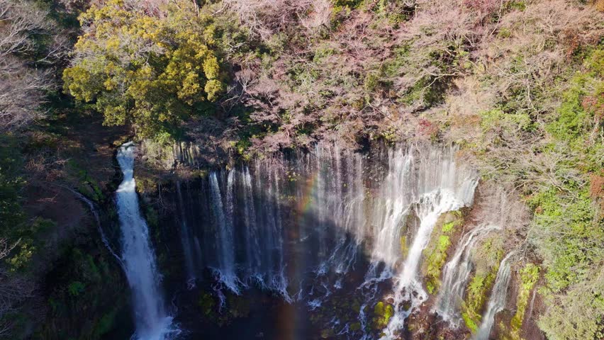 Stunning high angle aerial shot of a wide waterfall with a beautiful bright rainbow forming in the mist over the plunge pool.